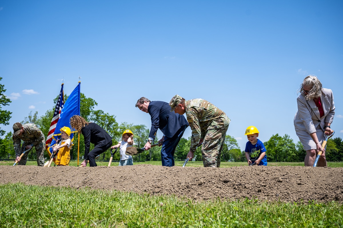 Five adults, military and civilian, shovel dirt with three children wearing hard hats