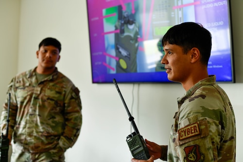 Two airmen stand by a television, with on holding a radio.