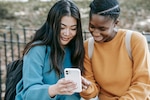 Two young women sit on a bench smiling and looking at a smartphone.