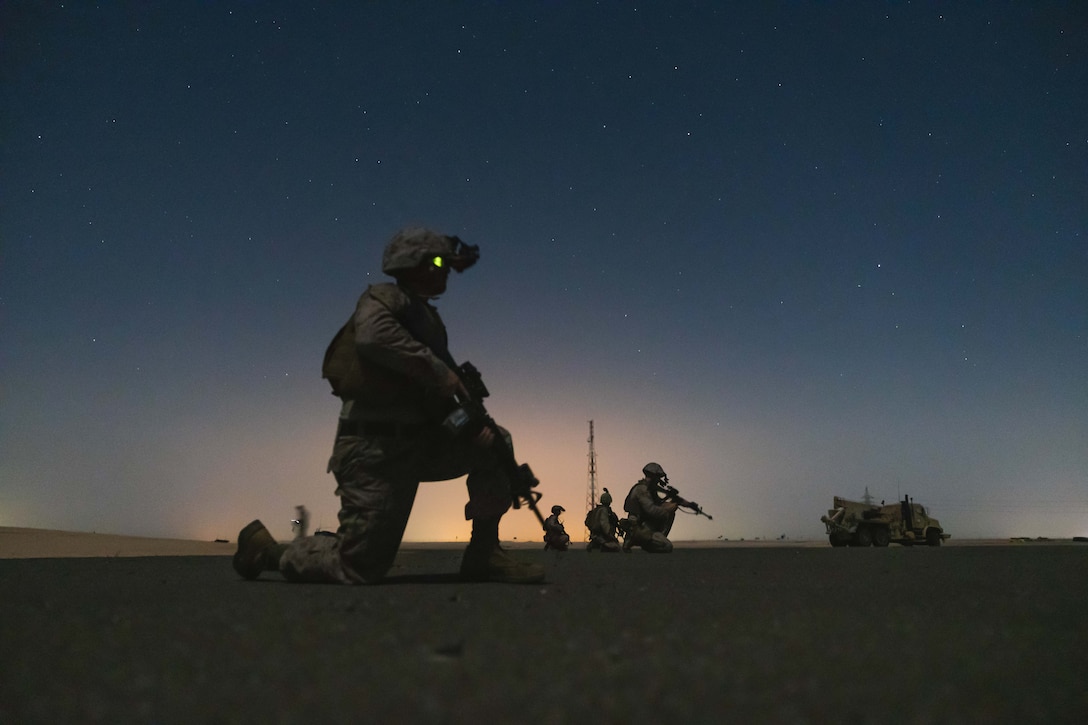 A group of Marines holding weapons kneel in a desert location in limited light as stars are seen in the sky. A vehicle sits on the right and a small tower is behind them.