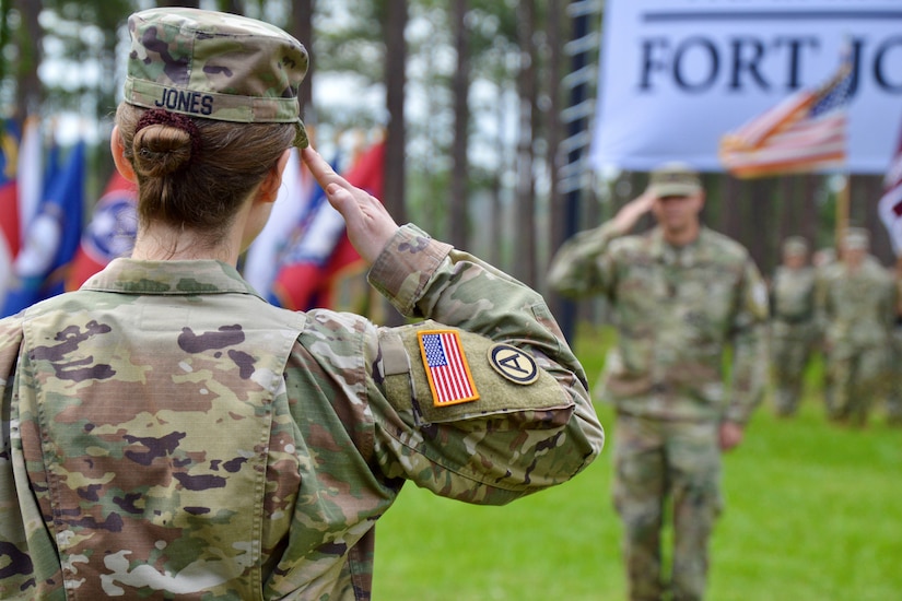 Two Army officers salute each other during a ceremony. A large sign and flags are seen in the background.