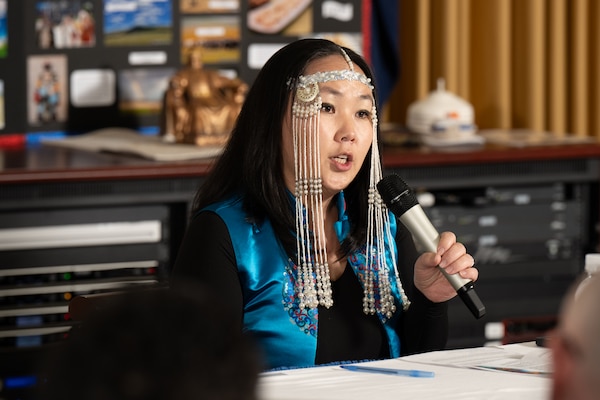 Nasa Primas speaks into a microphone while seated behind a table and wearing a draping Mongolian headband