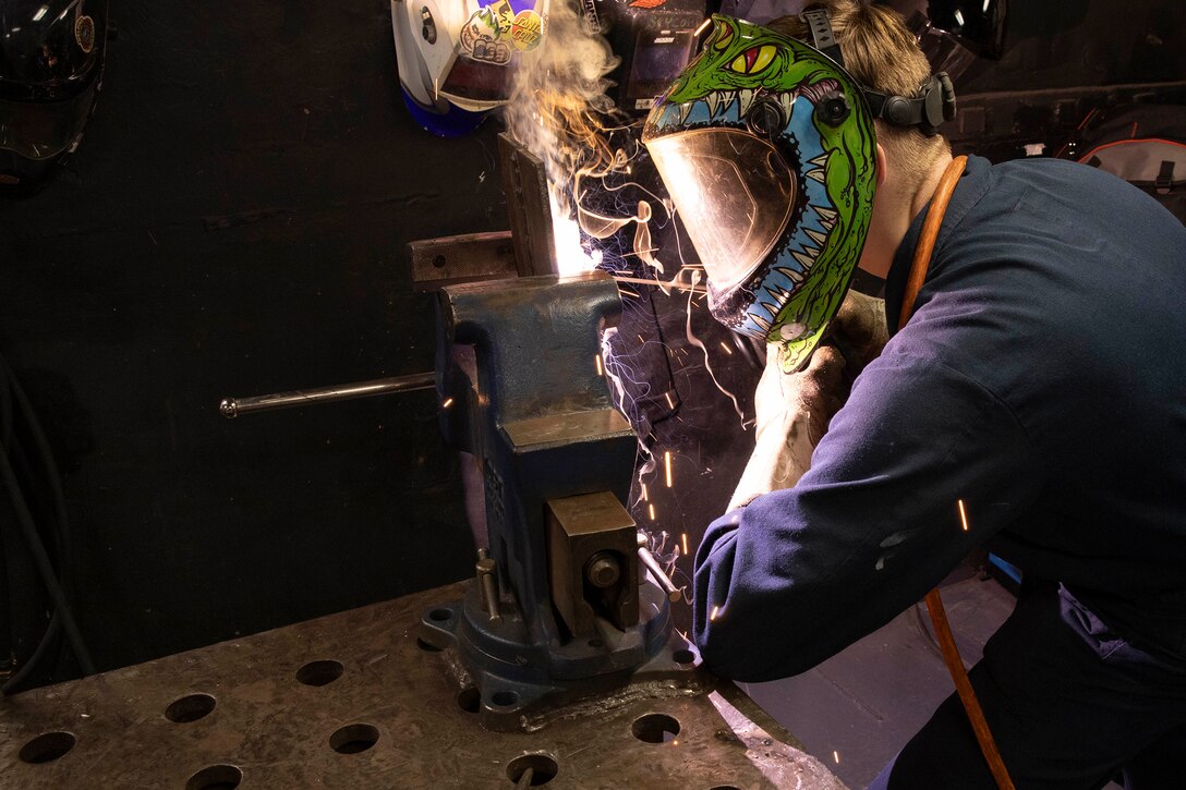 A sailor wears facial protection while welding metal inside a ship. Smoke can be seen rising from the welding tool.
