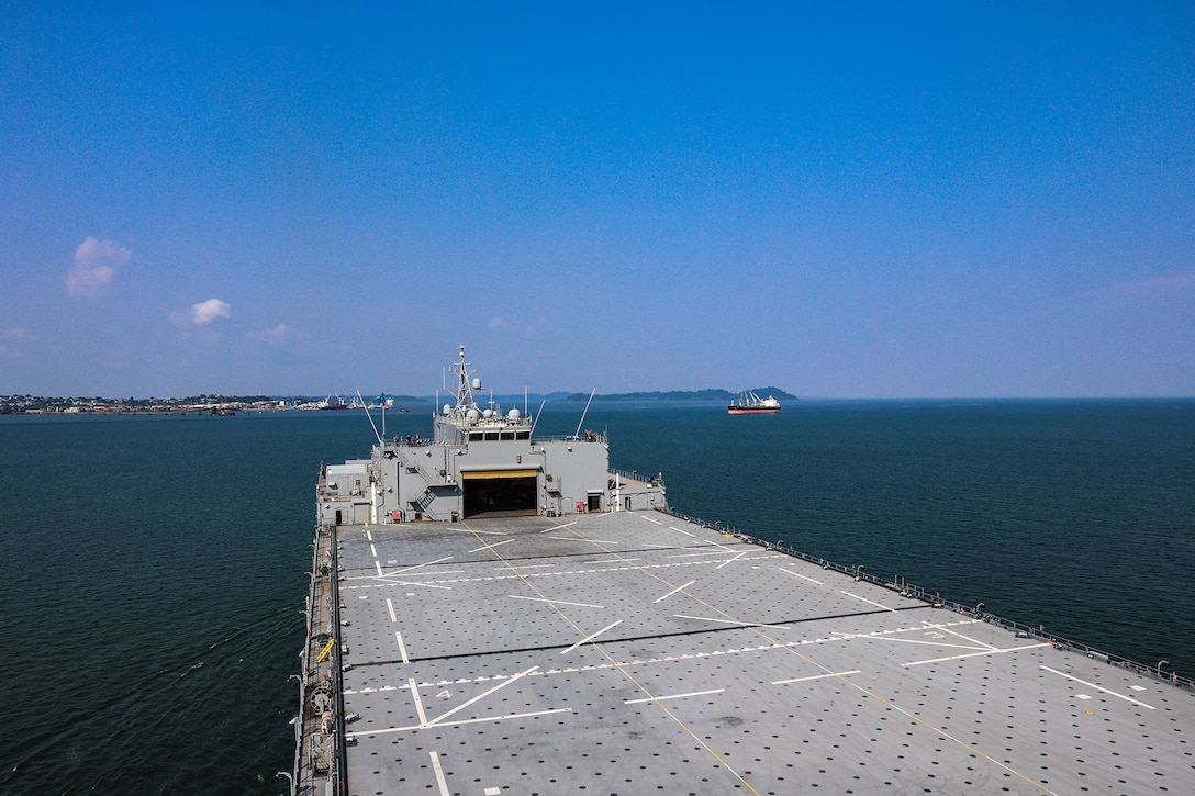 The bow of a ship in the ocean heads toward a port under a mostly clear sky with some clouds. A smaller ship is in the distance.
