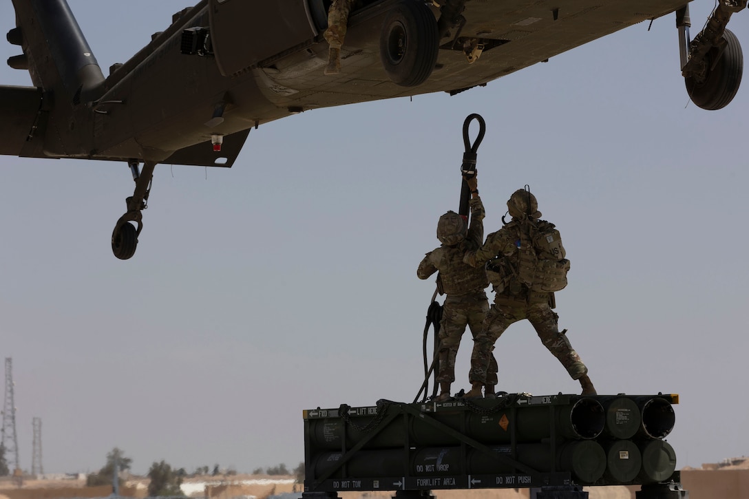 Soldiers reach up to attach a rope holding a rocket system pod to the underside of a military helicopter.