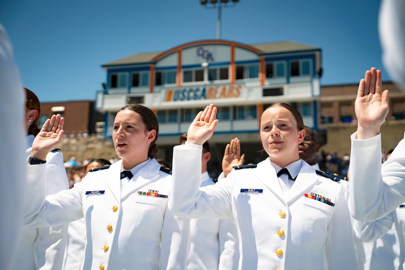 Coast Guard cadets in dress uniforms stand in formation while holding up their right hands with a building in the background.
