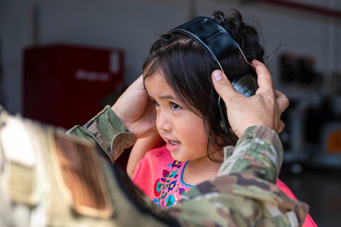A close-up of an airman’s hands putting headphones on a military child with a blurred shop in the background.