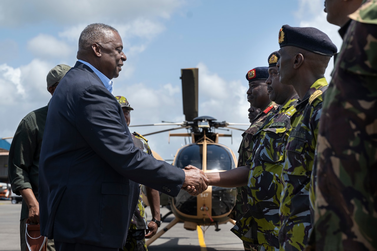 A man wearing civilian attire shakes hands with members of a foreign military in uniform.
