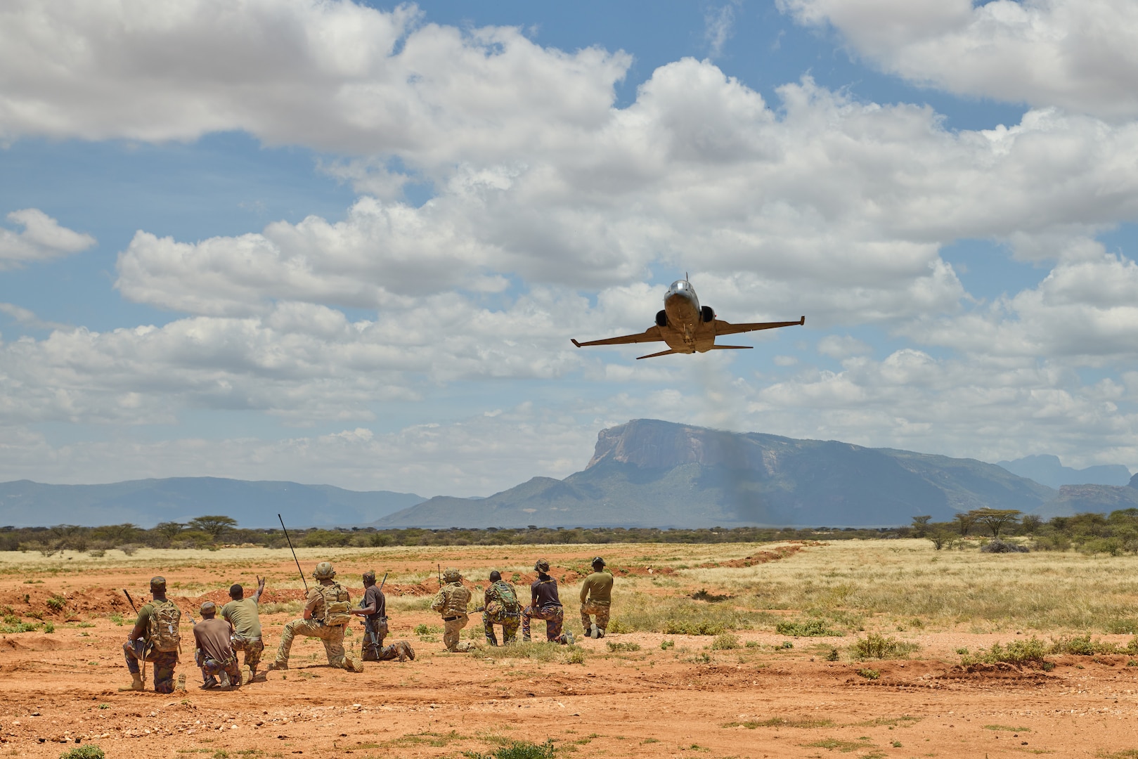 Service members in uniform kneel as a jet flies above them.