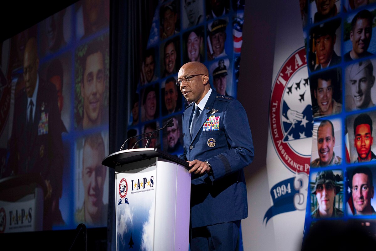An officer in a dress military uniform stands behind a lectern speaking into a microphone. The sign on the lectern says “TAPS - Tragedy Assistance Program for Survivors.” Behind the person are photos of the faces of fallen service members.