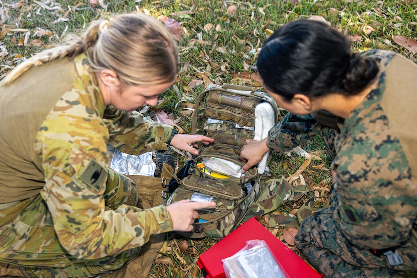 A U.S. sailor and an Australian soldier, seen from above, kneel over medical supplies in a grassy field.
