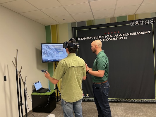 Two men stand in front of a display. One man is utilizing a virtual reality headset.