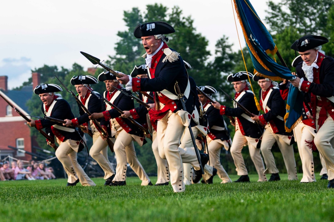 A group of soldiers in colonial uniforms run across a field in formation carrying rifles and a flag.