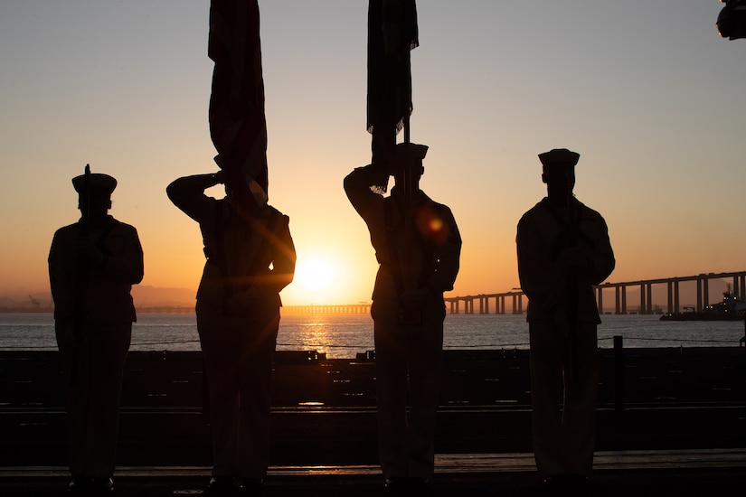 Four sailors present the colors aboard a Navy ship at twilight. They are shown in silhouette.
