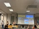 A woman holding a microphone stands next to a screen projecting a PowerPoint slide that reads "Environmental Justice Basics for Regulatory." A small crowd looks on.