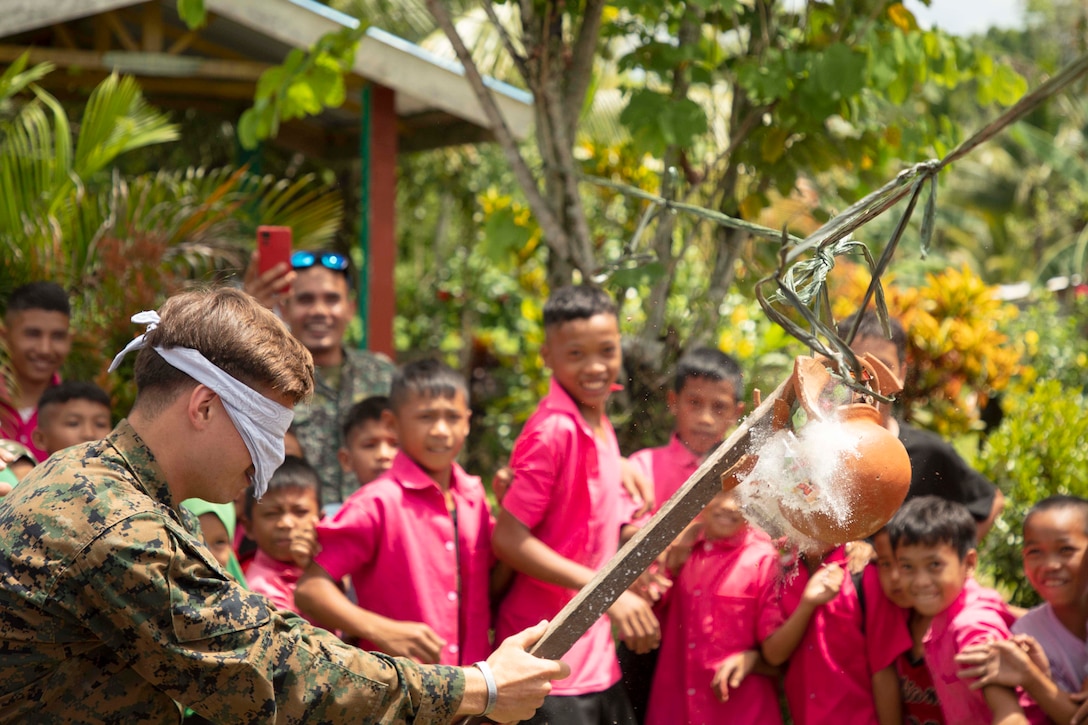 A Marine wearing a blindfold uses a stick to hit a fruit hanging from a tree as school students in uniforms laugh while watching surrounded by trees.