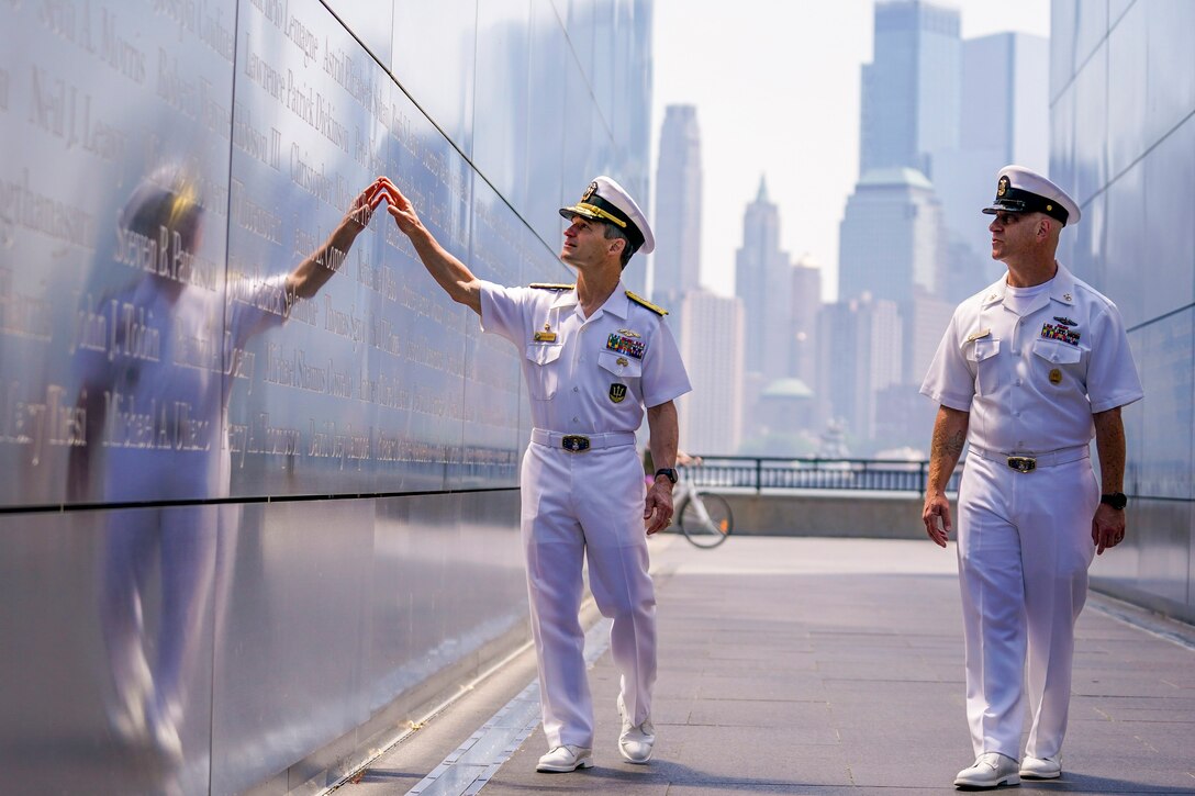 Two sailors walk while touching a memorial wall with tall buildings in the background.