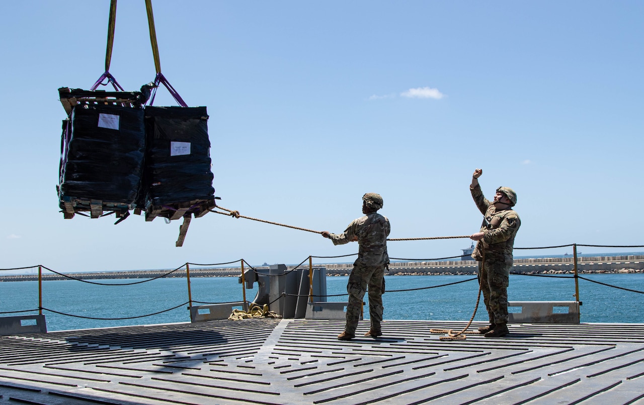 Two soldiers guide a pallet containing humanitarian assistance cargo onto a ship. The pallet is suspended by straps from a crane.