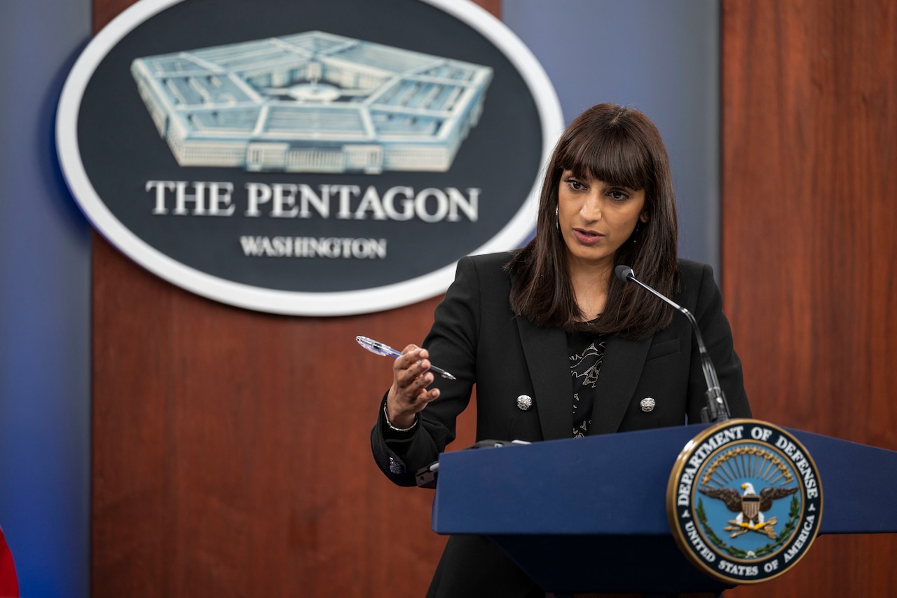 A woman at a lectern is gesturing while talking to people off screen. Behind her is a sign that indicates that she is at the Pentagon.