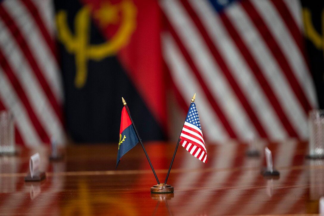 Mini American and Angolan flags sit on a table, with full-size flags from both nations in the background.