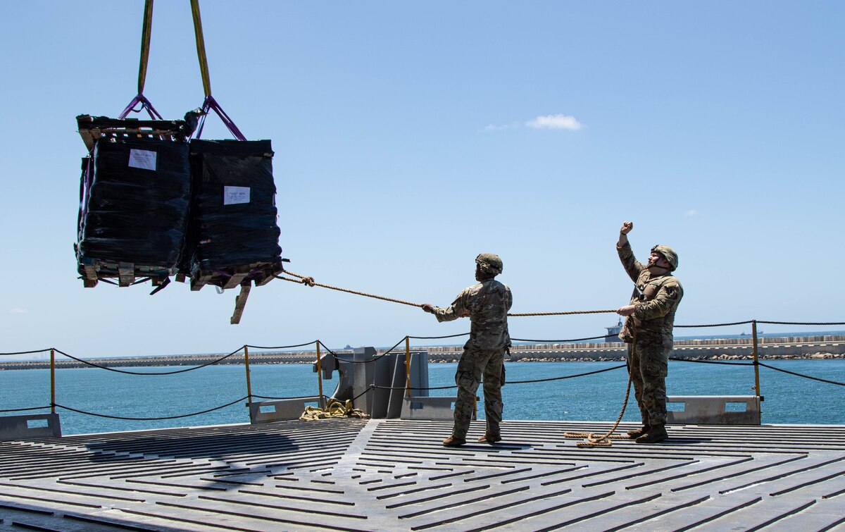 Two soldiers guide a pallet containing humanitarian assistance cargo onto a ship. The pallet is suspended by straps from a crane.