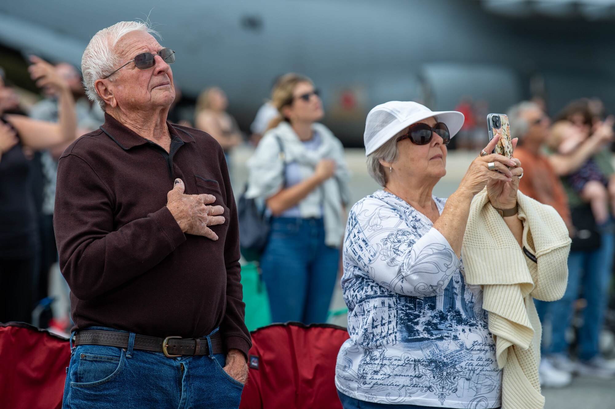 Spectators render respect and capture imagery of the U.S. Army Parachute Team, ‘The Golden Knights’ aerial performance during the National Anthem at the start of the 2024 First State Airshow at Dover Air Force Base, Delaware, May 19, 2024. The airshow was a free, two-day event, open to the public with 23 static displays and 18 aerial performers. (U.S. Air Force photo by Roland Balik)