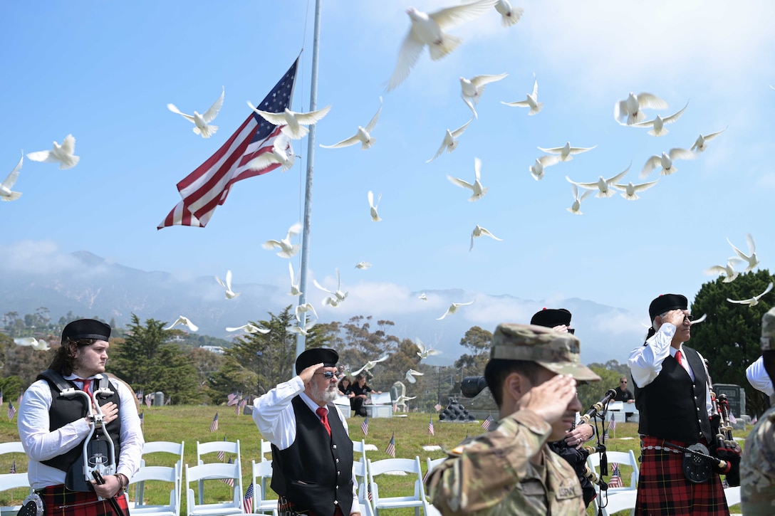 Service members and people wearing kilts salute as doves fly into the sky around a U.S. flag on a flagpole in a cemetery.