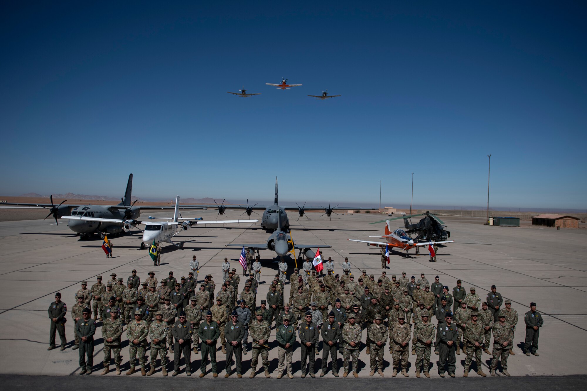 Representatives from the militaries of Peru, Colombia, Chile, Ecuador, Brazil, France and the United States pose for a group photo during the opening ceremony for Resolute Sentinel 2024 at Grupo 4 in La Joya, Peru, May 27, 2024, while military aircraft is shown behind and overhead.