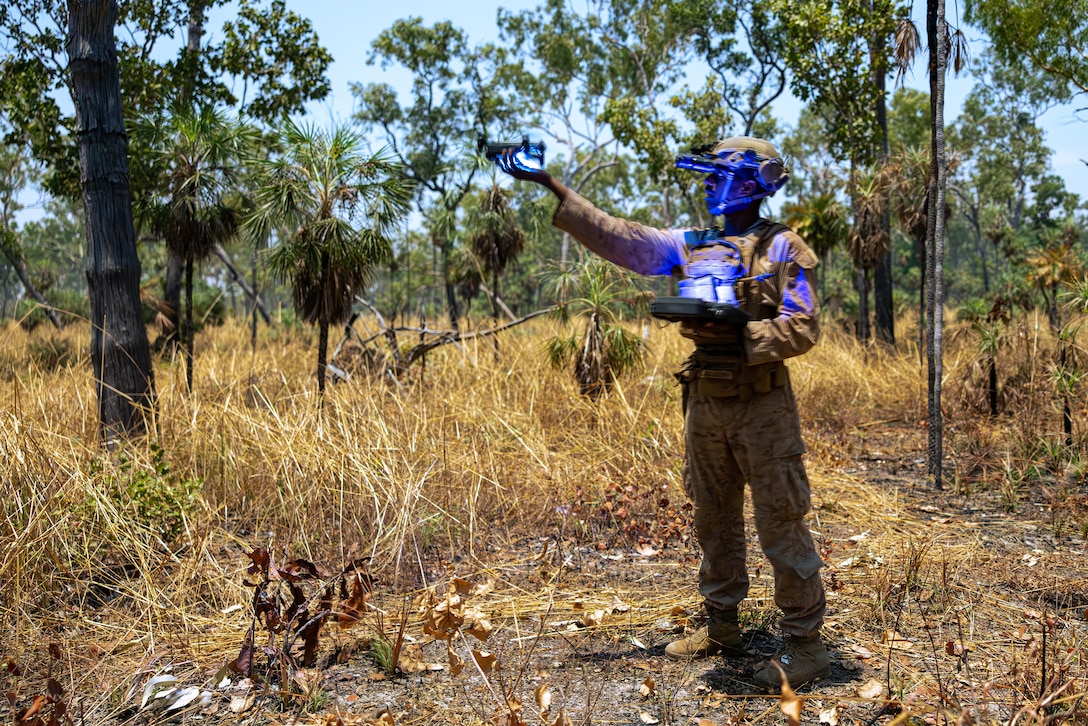 A uniformed Marine operates a small unmanned aircraft system in an outdoor training area. The device is emitting a blue light.