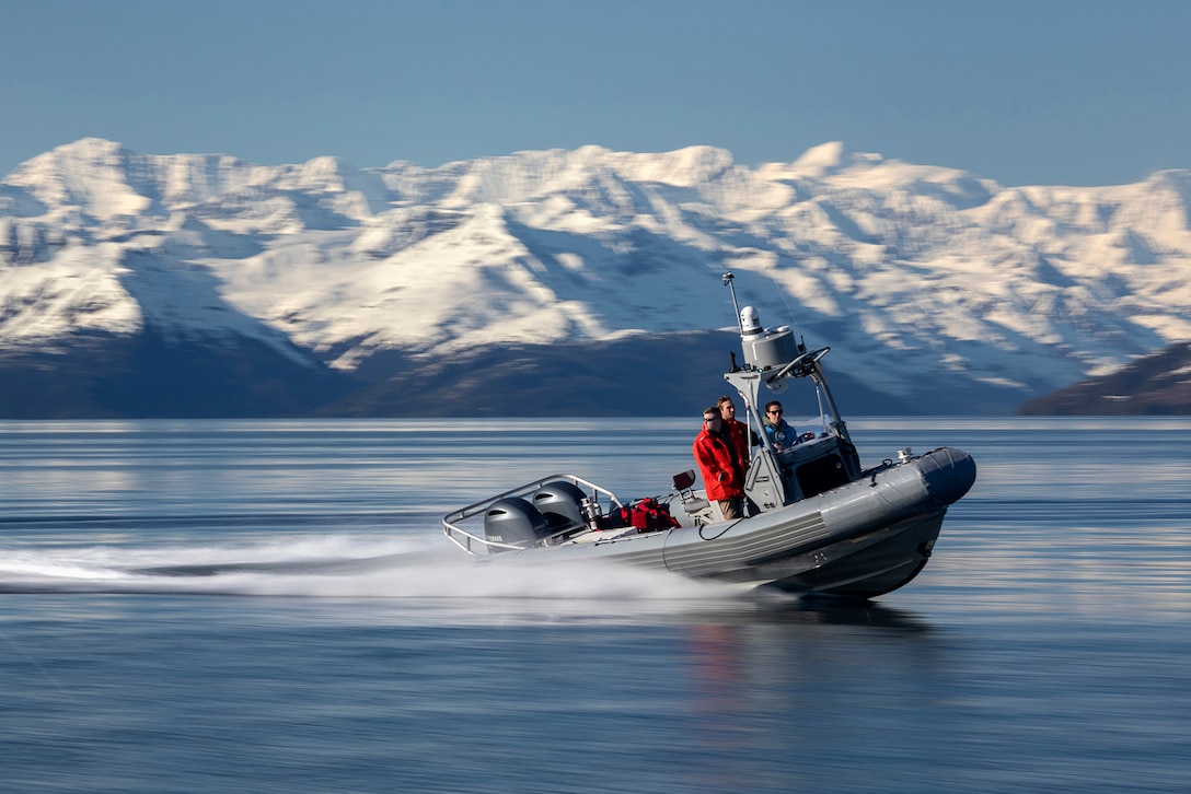 National Guard members ride in a small boat, leaving a wake behind them. Snow-capped mountains are visible behind the water.