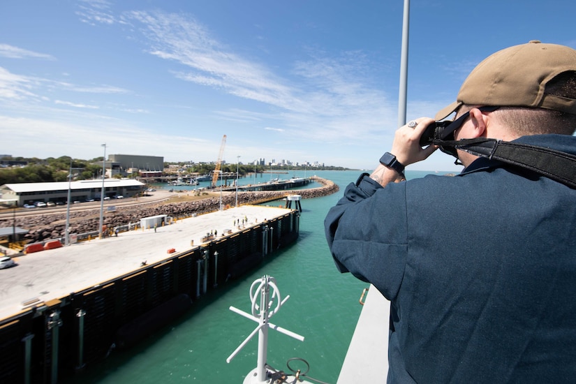 A sailor looks through binoculars as a ship arrives in port.
