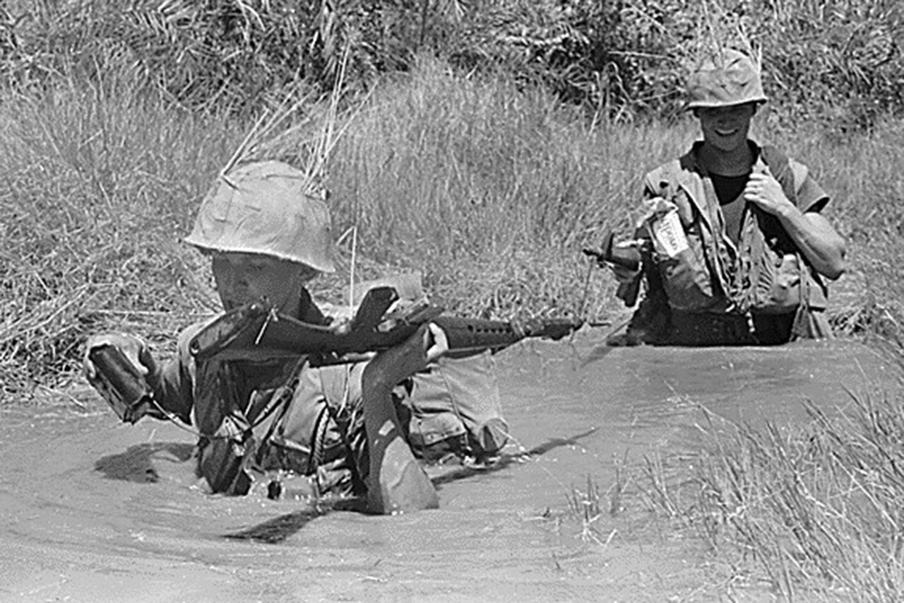 Two service members with rifles wade through chest-deep water.