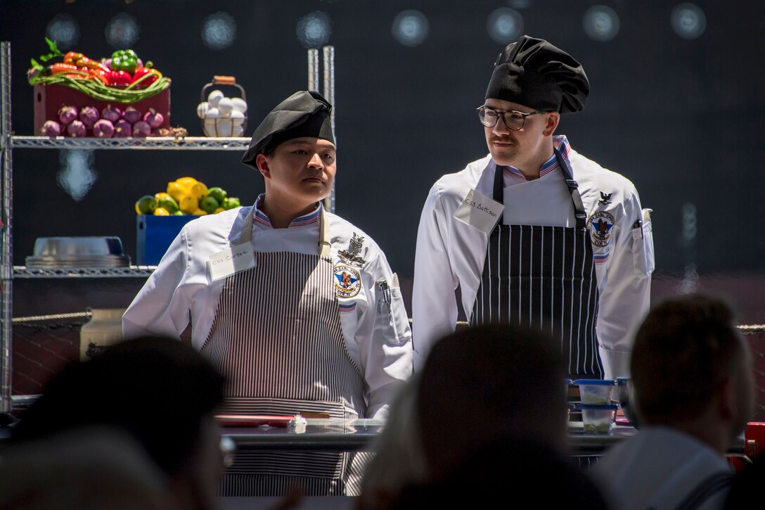 Two sailors in culinary gear stand under spotlights with shelves of food behind them in front of a seated audience.