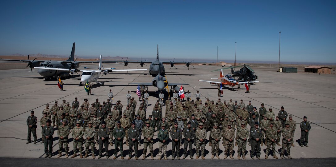 Representatives from the militaries of Peru, Colombia, Chile, Ecuador, Brazil, France and the United States pose for a group photo during the opening ceremony for Resolute Sentinel 2024 at Grupo 4 in La Joya, Peru, May 27, 2024, while military aircraft is shown behind and overhead.
