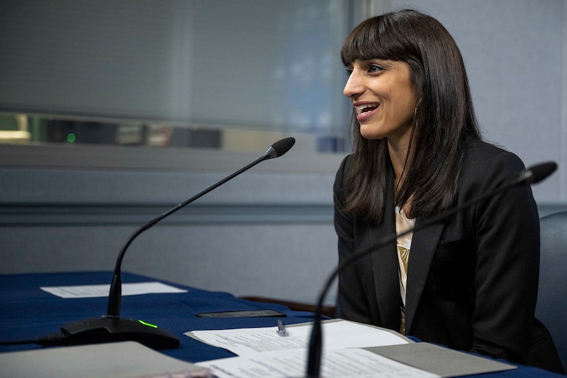 A person in civilian attire sits at a table with a microphone.