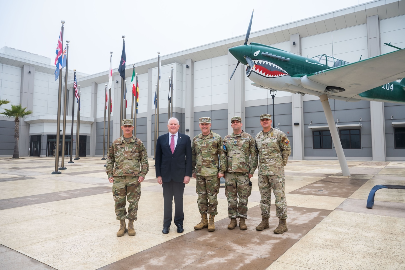 Five gentlemen, four in military uniform and on in a suit, stand together for a photo outside a building with an aircraft static display.