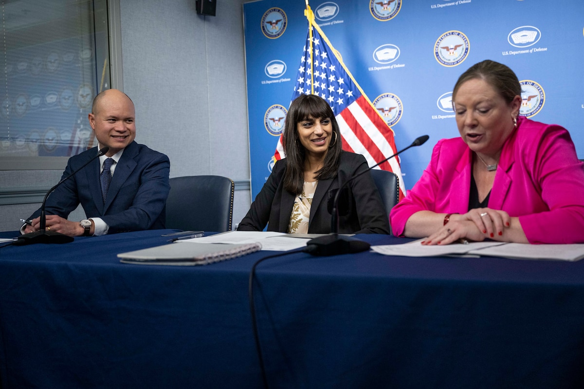 Three people in civilian attire sit at a table with a United States flag in the background.