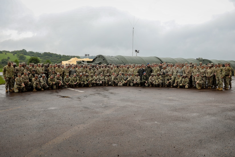 A group of soldiers poses for a photo outside during an overcast day in front of tent buildings.