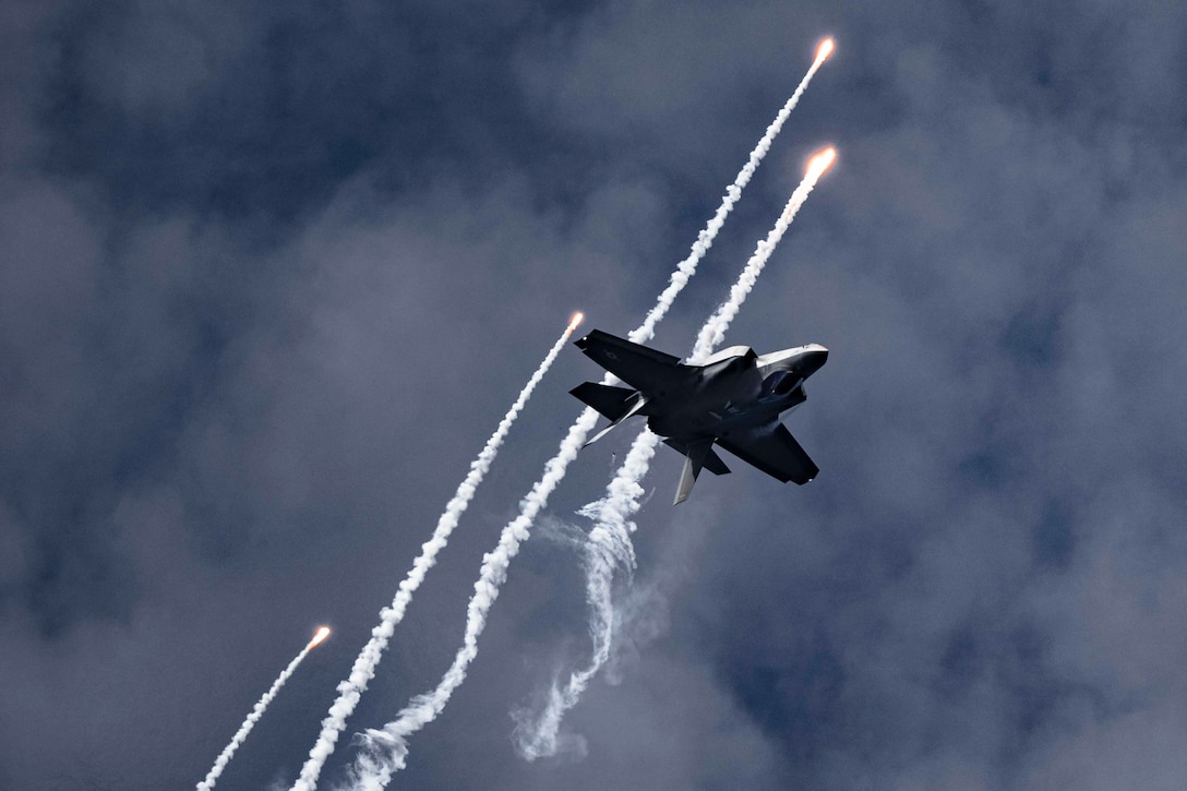 A military jet flies upside down through the air as flares leave smoke trails behind it.