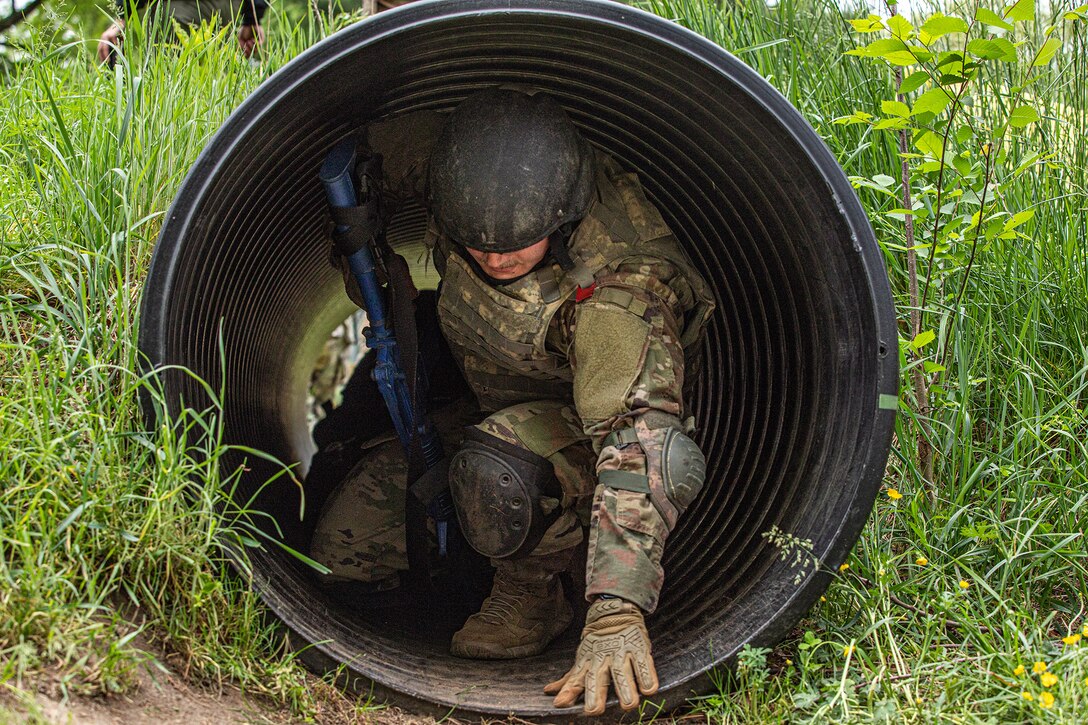 A uniformed soldier with a weapon exits a tunnel in a grassy area during daylight training.