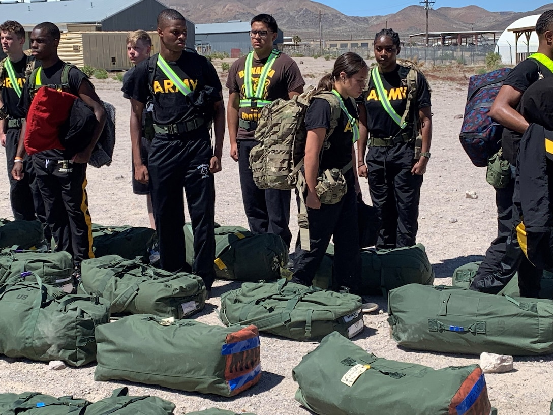 JORTC members stand in a line at attention in front of green duffel bags on the ground. The students are wearing black t shirts with the word ARMY on them. Two students are not in line and look as if they are walking along the line inspecting their fellow JROTC members.