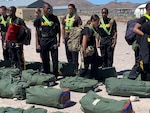 JORTC members stand in a line at attention in front of green duffel bags on the ground. The students are wearing black t shirts with the word ARMY on them. Two students are not in line and look as if they are walking along the line inspecting their fellow JROTC members.