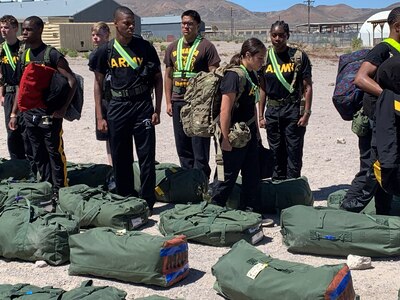 JORTC members stand in a line at attention in front of green duffel bags on the ground. The students are wearing black t shirts with the word ARMY on them. Two students are not in line and look as if they are walking along the line inspecting their fellow JROTC members.