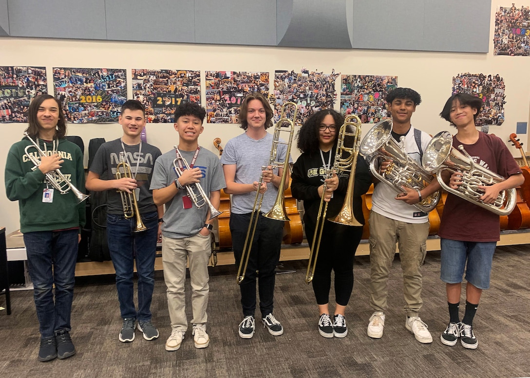 7 high school age teen agers stand in a line facing the camera all holding a musical instruments. three are cornets, two are trombones the last two are Tubas.