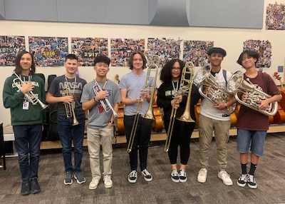 7 high school age teen agers stand in a line facing the camera all holding a musical instruments. three are cornets, two are trombones the last two are Tubas.