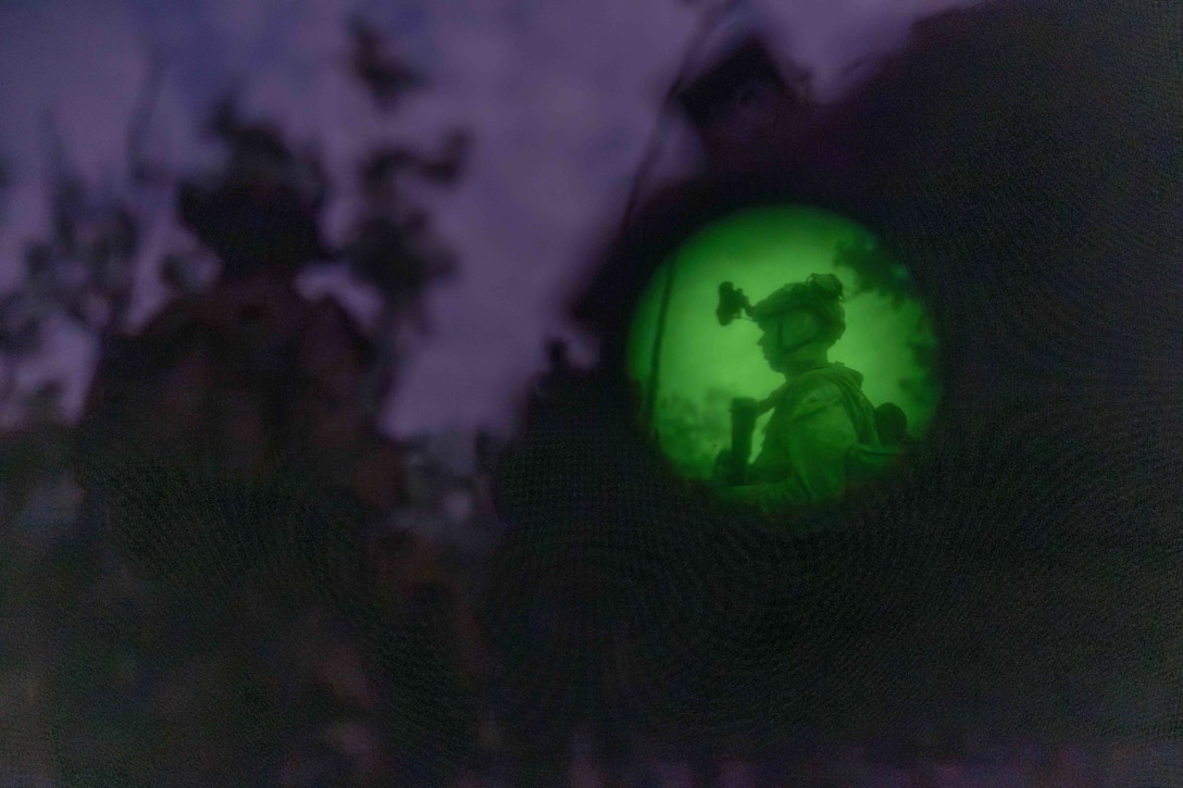 A Marine stands in a forest as seen in a night vision lens to the right as fellow Marines gather nearby.