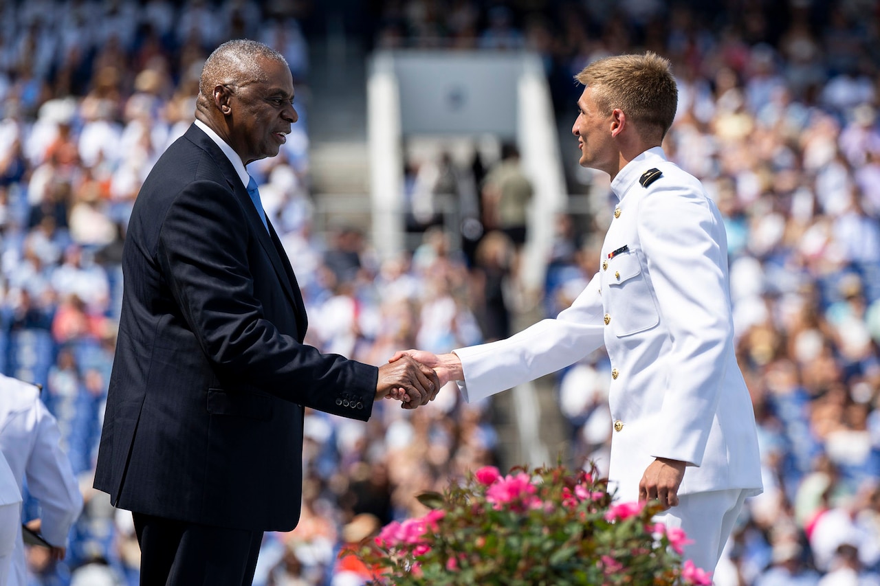 Secretary of Defense Lloyd J. Austin III shakes hands with a sailor in dress uniform in a stadium full of people.