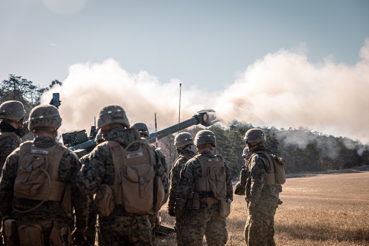 Seven Marines in camouflage uniforms, field vests and helmets are standing outside in a field with their backs to the camera, looking at a cannon-shaped gun with smoke coming out of the barrel