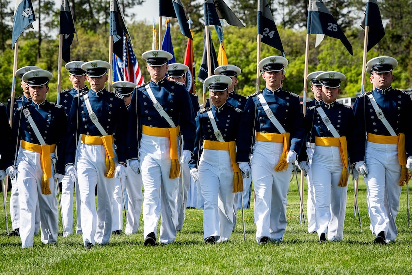 U.S. Air Force Academy cadets, some holding flags, walk in formation on a grass field.