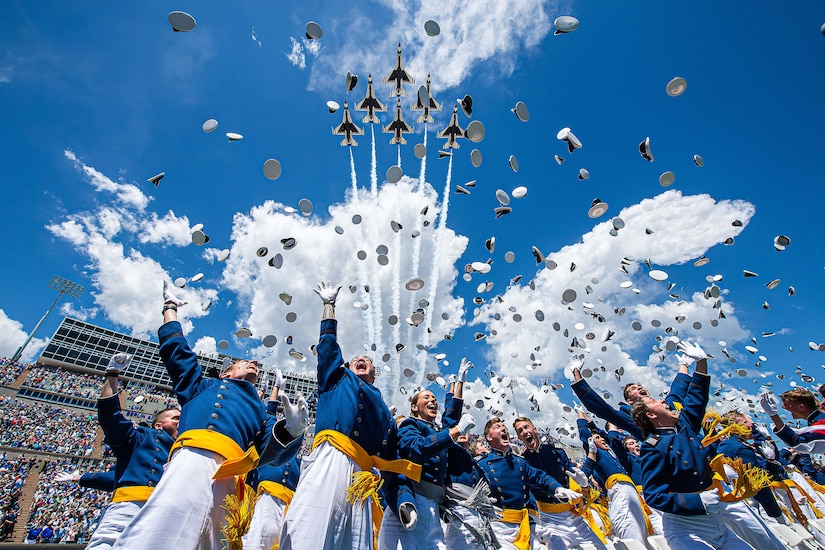 Air Force cadets toss caps in the air in a stadium as jets fly overhead.